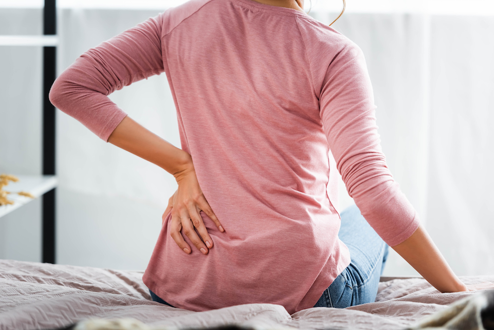 Back view of a woman sitting on a bed, experiencing pain in her tailbone area